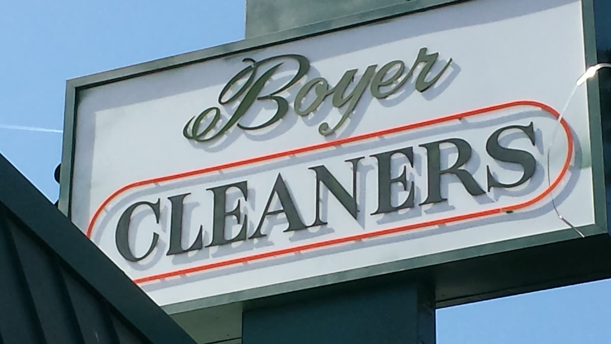 The Boyer Cleaners blade sign above the sidewalk: script Boyer over serif CLEANERS, ringed by an orange neon tube.